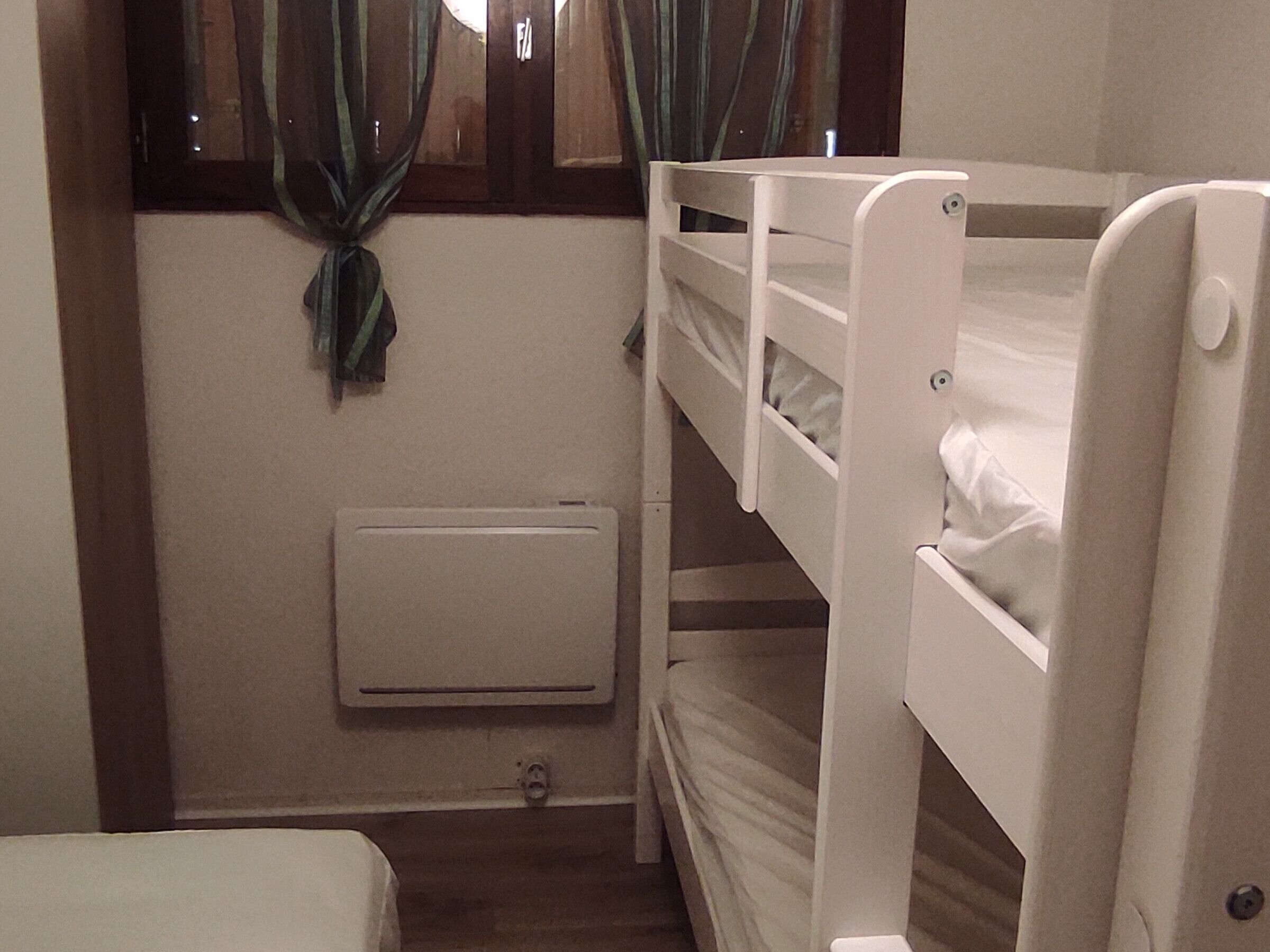 A bunk bed room with a window, curtains, a radiator, and a small bedside table.