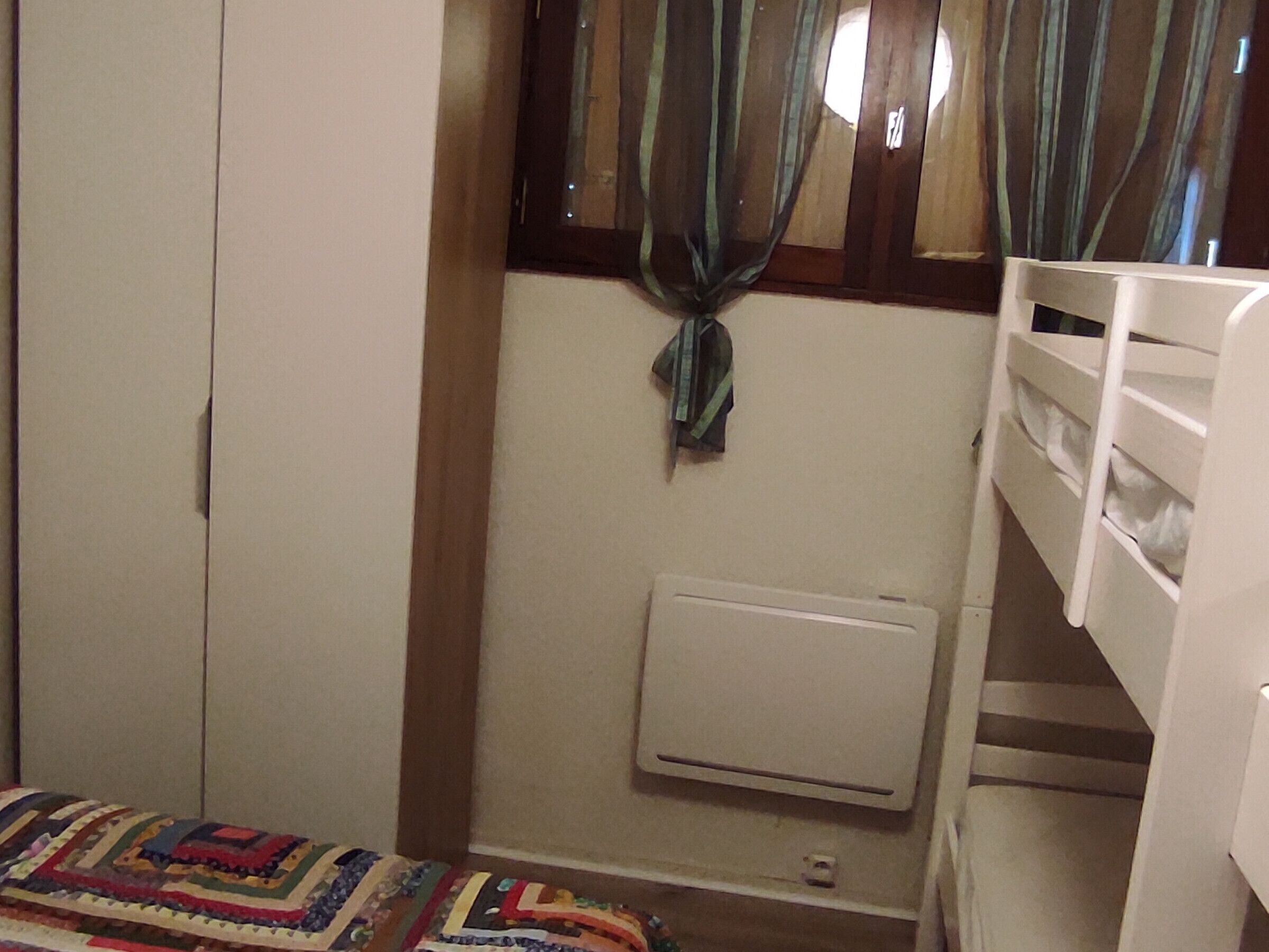 A bedroom with a bunk bed, a wardrobe, a radiator, and a window with curtains.