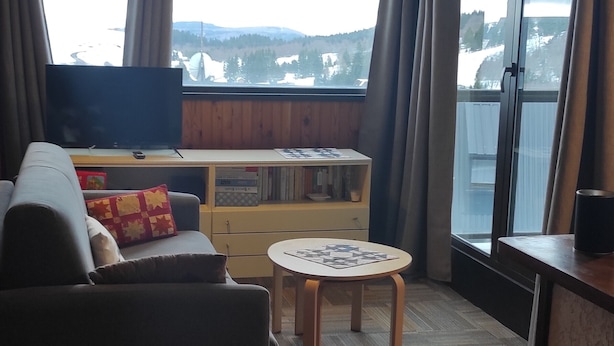 Una sala de estar con sofá, una mesita, televisor y ventana con vistas a montañas.