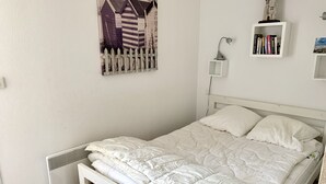 En snyggt bäddad säng i ett rum med en vit vägg, en liten hylla med böcker och en tavla som föreställer ett hus vid stranden.