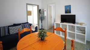 Una sala de estar con un sofá, una mesa de madera con una planta, un televisor sobre un mueble bajo y un ventilador.