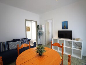 Ein Wohnzimmer mit einer Couch, einem Holztisch mit Pflanze, einem Fernseher auf einem Schrank und einem Ventilator.