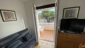 Ein Zimmer mit einer Couch, einem Fernseher auf einer Holzkommode und einer Tür zu einem Außenbereich mit einem weißen Stuhl und einem Baum.