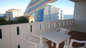 Ein Balkon mit einem weißen Tisch und Stühlen, einem gestreiften Sonnenschirm und Blick auf Gebäude sowie das Meer.