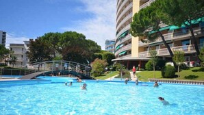 Piscina com pessoas nadando e passeando. Árvores e um prédio com varandas ao fundo.