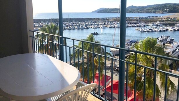 Un balcón con mesa y sillas, con vista a un puerto deportivo lleno de barcos y una vista despejada de la costa.