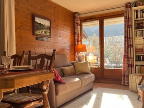Ein gemütliches Wohnzimmer mit Holzwand, einer Couch, einem Esstisch mit Stühlen, einem Bücherregal und einem Fenster mit Vorhängen.