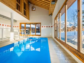 A large indoor swimming pool with clear glass walls, wooden beams, and a view of a snowy landscape. - Charming 3-Room South, Pool & Pets Allowed (Samoëns)