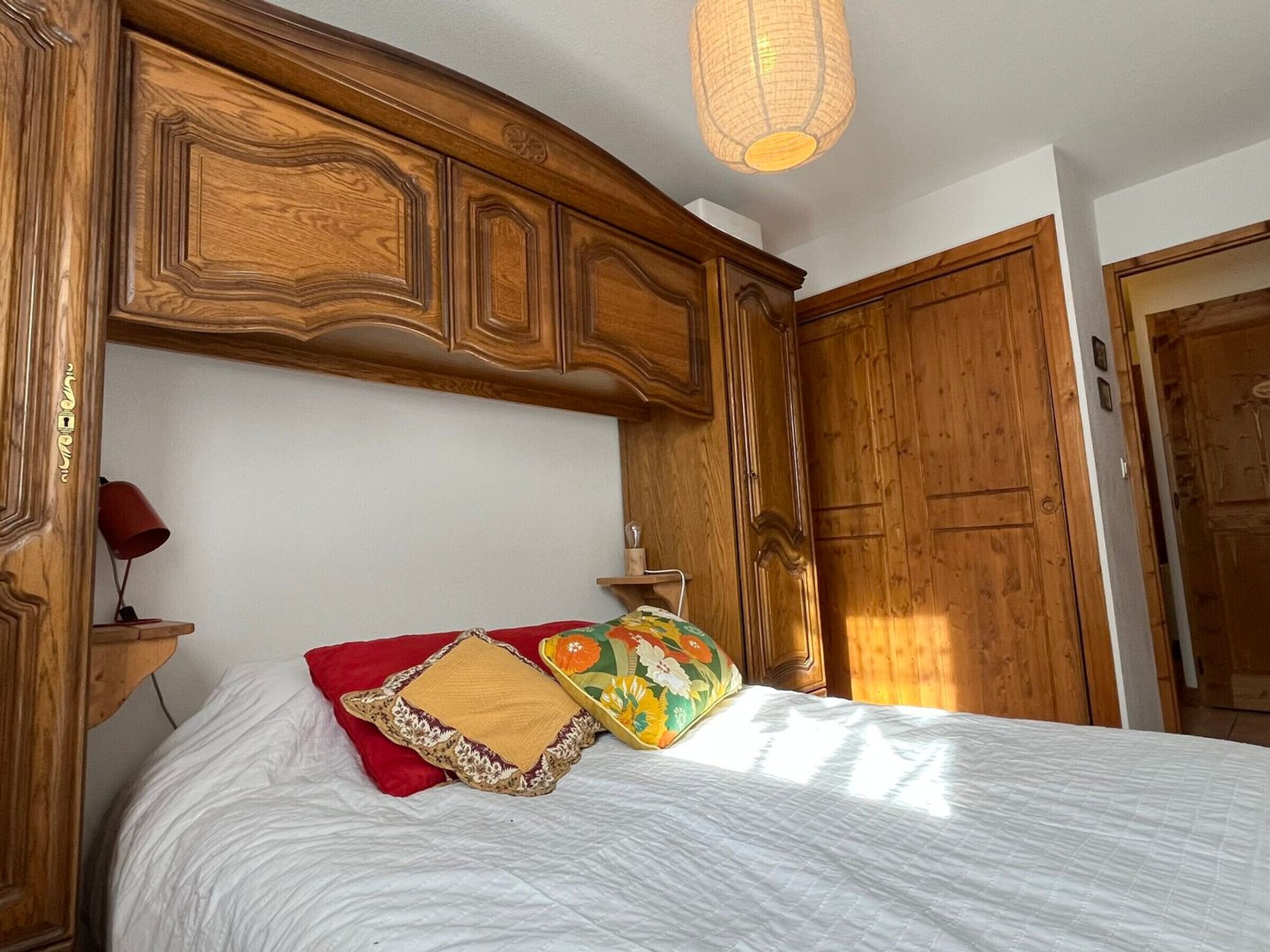 A bedroom with a wooden headboard, a bed with white bedding and multiple pillows, a red lamp on a bedside table, and a wooden wardrobe.