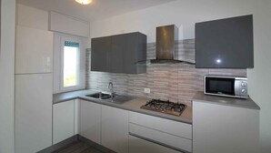 A modern kitchen with white cabinets, a stainless steel range hood, a microwave, and a window with blinds. - Holiday apartment mit Pool und Meerblick (Lignano Sabbiadoro)