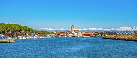A waterfront town with a prominent tower, boats docked along the shore, and a backdrop of snow-capped mountains.