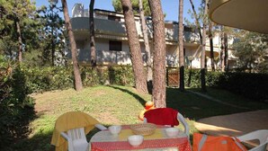 An outdoor dining area with a table set for four, surrounded by trees and a building in the background. - Holiday apartment mit Terrasse (Lignano Sabbiadoro)