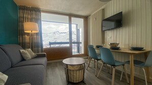 A modern living room with a sofa, a round coffee table, and a dining area with chairs and a table. A TV is mounted on the wall, and there is a large window with a view of snow-capped mountains. - Studio for 4 in Tignes, Pets allowed (Tignes)