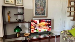 Un salón con una televisión mostrando una comida, un sofá con un cojín estampado y una estantería decorativa con diversos objetos.
