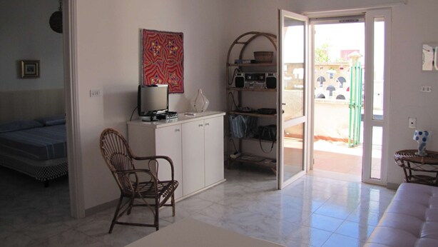 Ein Zimmer mit einem Bett, einem Schreibtisch mit Computer, einem Stuhl, einer zur Außenwelt führenden Tür und einer Wand mit einem farbenfrohen Kunstwerk.