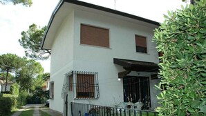 A two-story house with a white exterior, a covered porch, and a small garden area. - Holiday apartment mit Garten (Lignano Sabbiadoro)