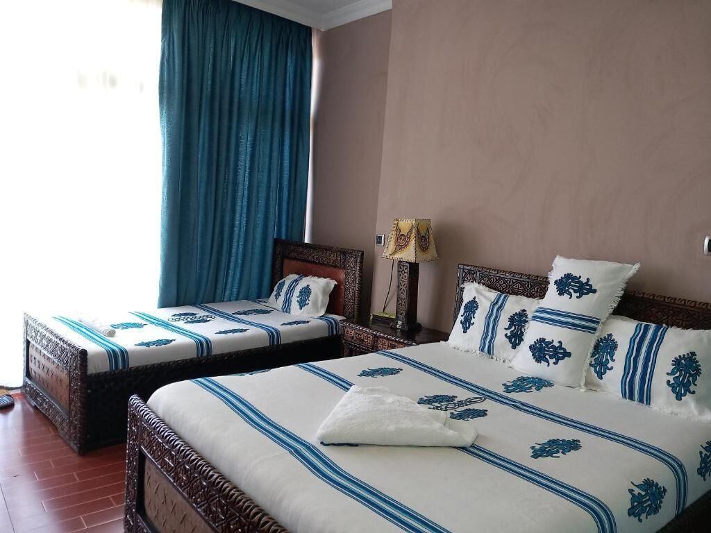 A hotel room with two beds, each with a patterned bedspread and pillows, a bedside lamp, and a window with blue curtains.