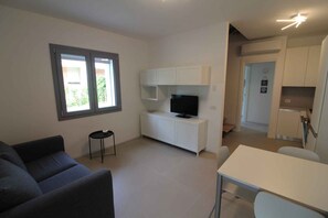A modern living room with a sofa, a TV on a white cabinet, and a dining area with a table and chairs. - Holiday apartment mit Balkon (Lignano Sabbiadoro)
