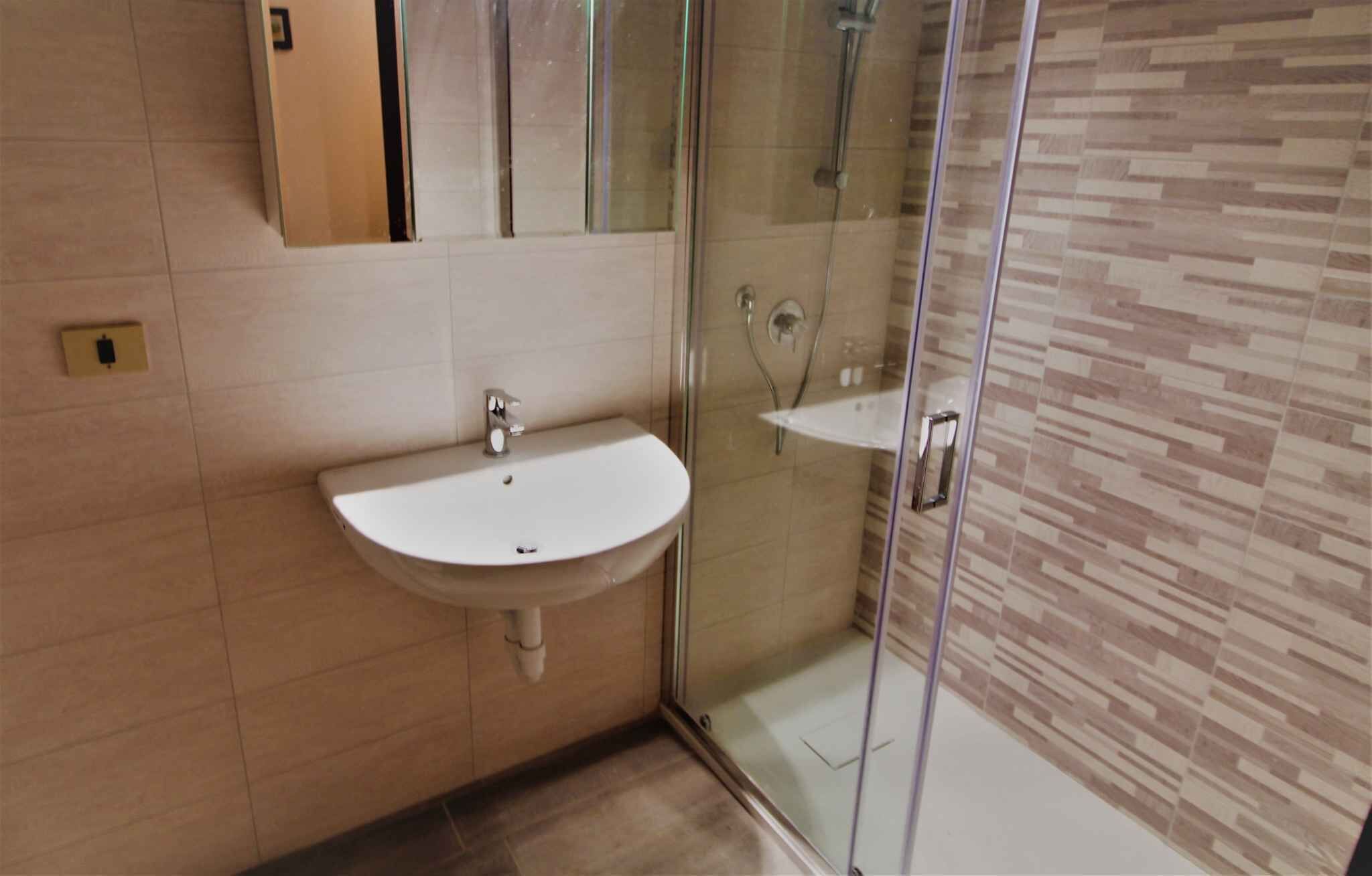 Baño con lavamanos blanco, cabina de ducha con cerramiento de vidrio y pared revestida con un diseño que imita la madera.