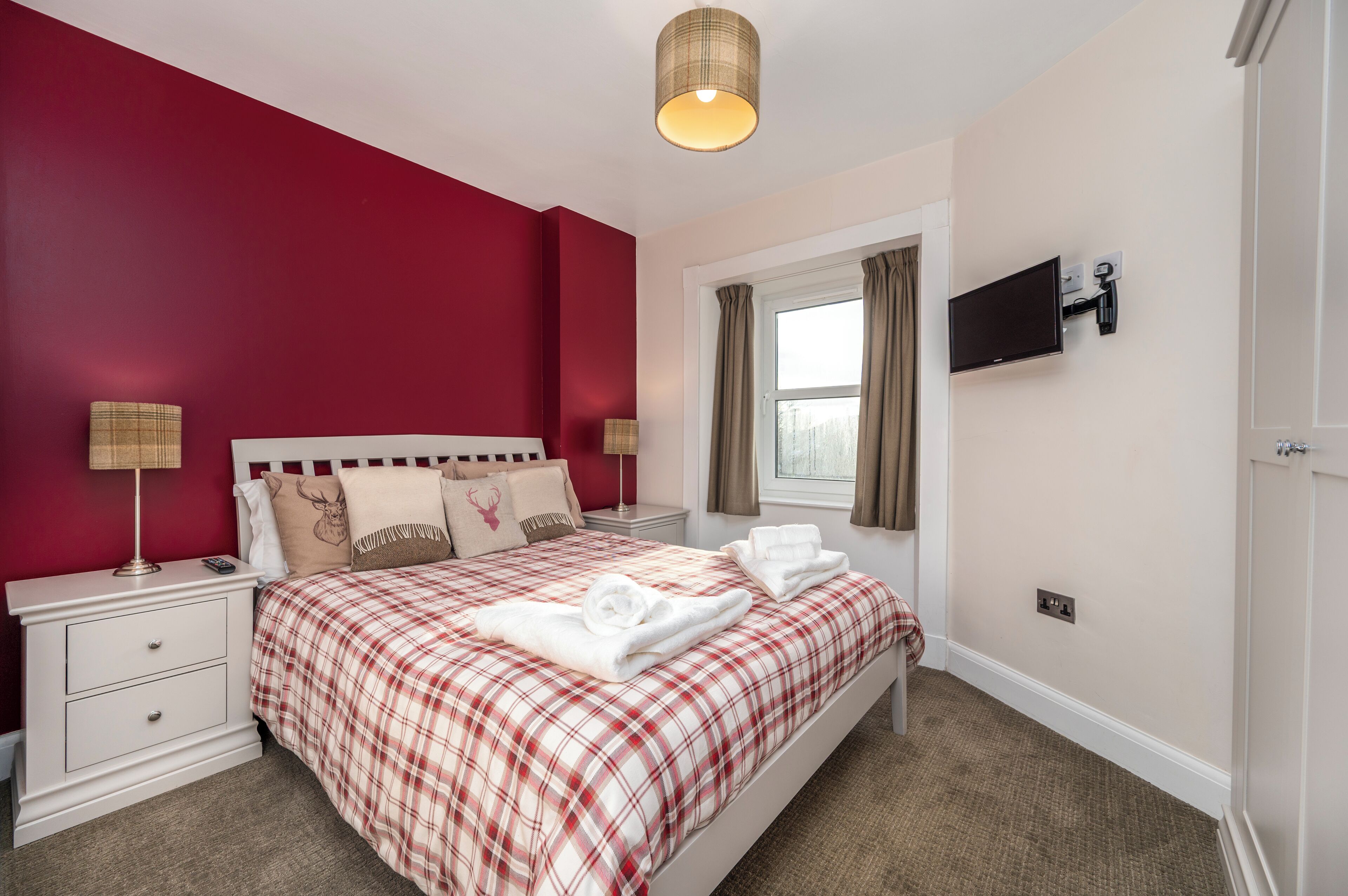 A neatly arranged bedroom with a red and white checkered bedspread, a white nightstand with a lamp, and a television mounted on the wall.