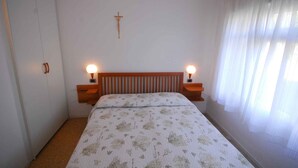 A bedroom with a wooden headboard, two bedside lamps, a cross on the wall, and a window with sheer curtains. - Holiday apartment mit Terrasse (Lignano Sabbiadoro)