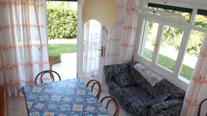 A dining area with a table covered in a patterned tablecloth, a sofa with a floral design, and large windows with curtains. - Holiday apartment mit Terrasse (Lignano Sabbiadoro)