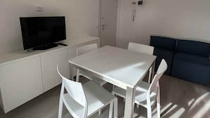 Una sala de estar minimalista con una mesa blanca, cuatro sillas, un sofá oscuro, un televisor sobre un mueble blanco y una puerta con interruptor de luz.