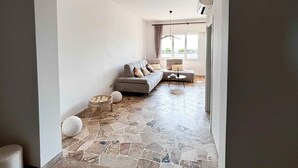 A spacious living area with a stone floor, a grey sofa with cushions, a small round table, and a hanging light fixture. - Holiday apartment with air conditioning (Jesolo)