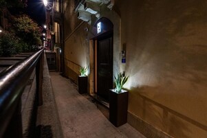 Vista nocturna del exterior de un edificio con barandilla metálica, una puerta con un letrero encima y dos plantas en macetas en la acera.