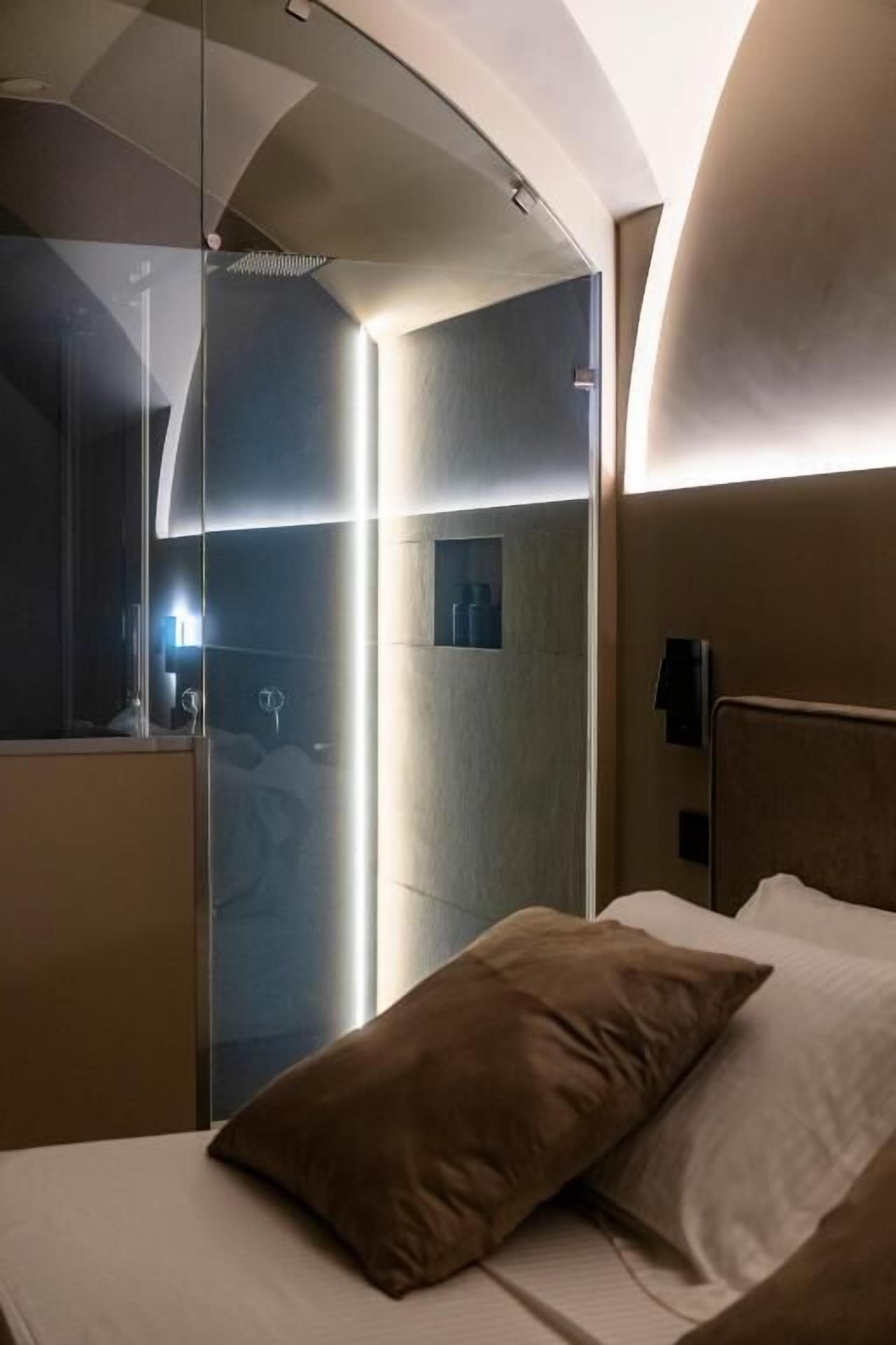 Una habitación de hotel moderna con techo curvo, una cama con dos almohadas, una mesita de noche y una mampara de vidrio que separa la habitación del baño.