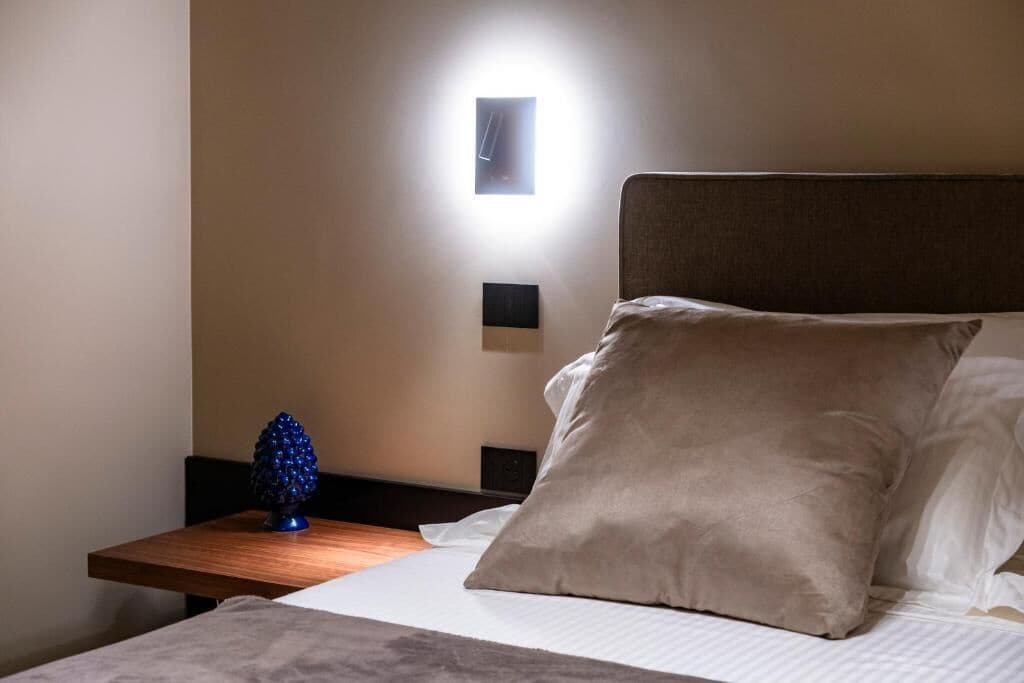 Una cama bien hecha con un almohadón beige grande, una mesita de noche con un objeto decorativo azul, y una lámpara de pared.