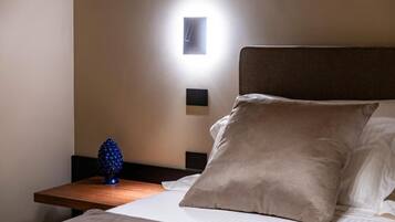Un lit bien fait, agrémenté d’un grand oreiller beige, d’une petite table de chevet sur laquelle repose un objet décoratif bleu, et d’un luminaire fixé au mur.