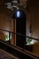 Una entrada arqueada a un edificio con una pantalla digital encima de la puerta, una planta en maceta a cada lado y una barandilla metálica en primer plano.