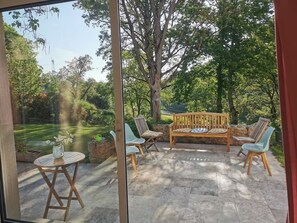 Une terrasse extérieure avec un banc et des chaises en bois, une petite table ronde ornée d’un vase et un sol en pierre.