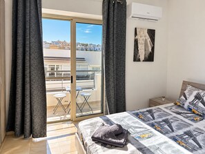 A bedroom with a bed, a nightstand, a potted plant, and a view of a cityscape through a sliding door. - Chic & Central 3-Bedroom Apartment in Gzira (Il-Gżira)