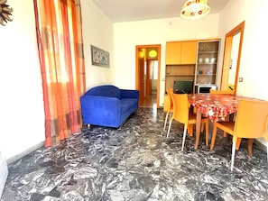 Un salon avec un canapé bleu, une table à manger avec des chaises orange et un coin cuisine équipé de placards et d’un téléviseur.