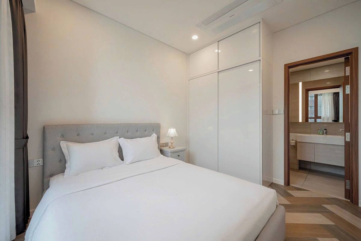 Una habitación de hotel moderna con una cama grande, una mesita de noche con lámpara, un armario y un baño visible a través de una puerta abierta.