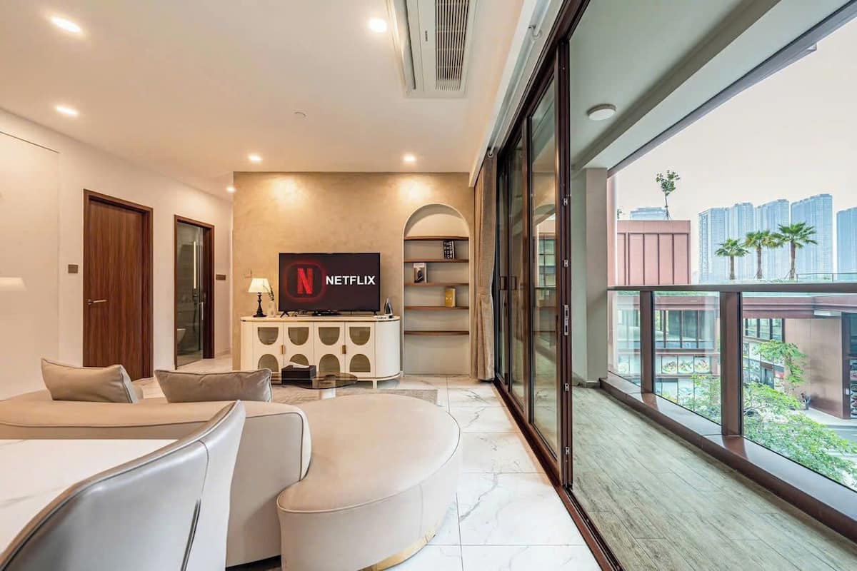 Una sala de estar moderna con un televisor de pantalla plana, un sofá y una mesa de centro. Hay un balcón con vistas a edificios y palmeras.