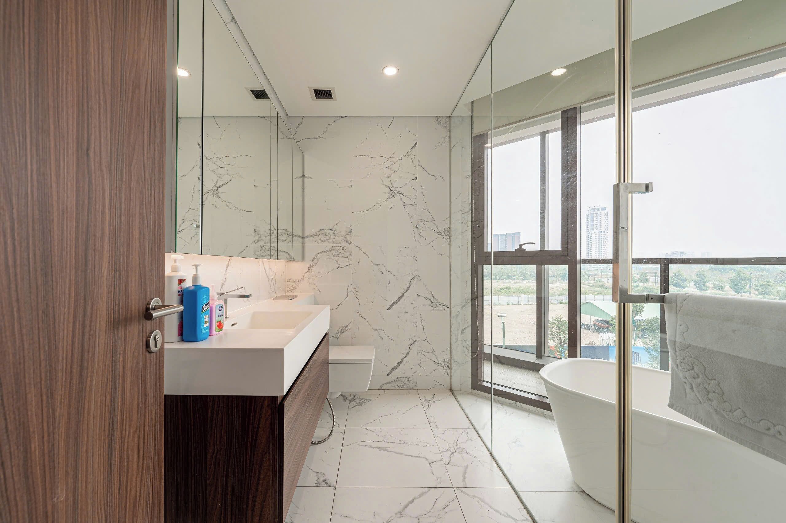 A modern bathroom with a marble wall, a large bathtub, and a view of the city through a large window.