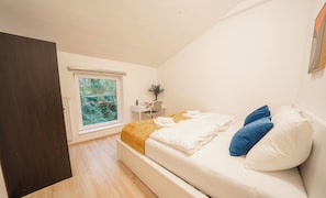 A bedroom with a bed, pillows, a desk, a chair, a window with a view of greenery, and a door. - 2 Bedroom Apartments 18 Min to center (Vienna)