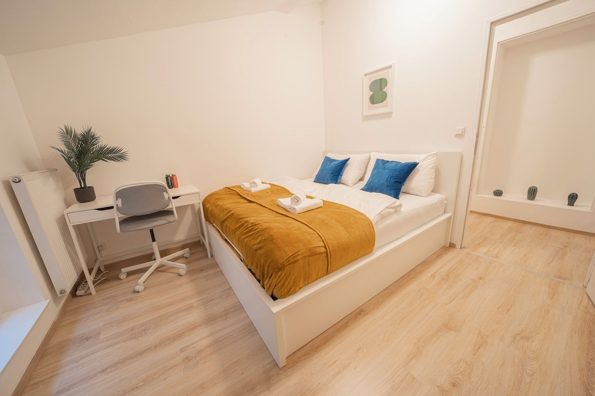A bedroom with a bed, a desk, a chair, a potted plant, and a wall-mounted shelf.