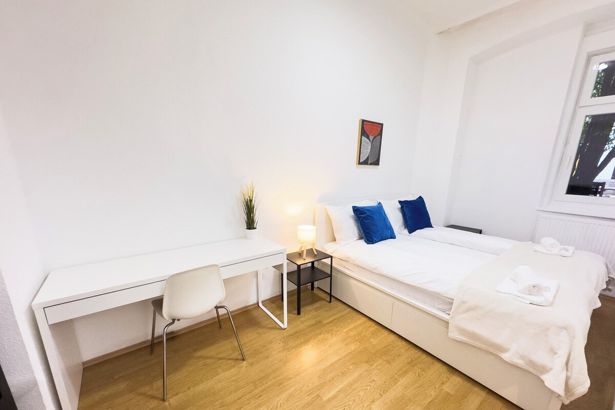 A modern, minimalist room with a white desk, a single chair, a bed with blue pillows, and a small bedside table with a lamp.