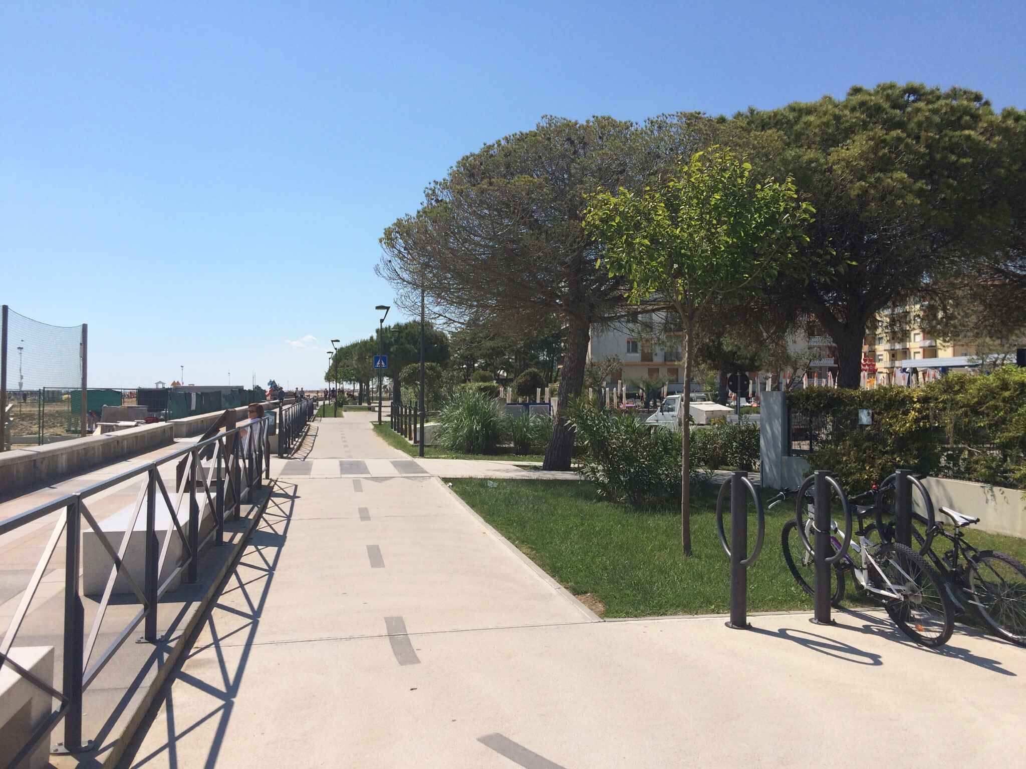 Un trottoir pour piétons agrémenté d’une rambarde métallique, d’arbres et d’un espace de stationnement pour vélos.