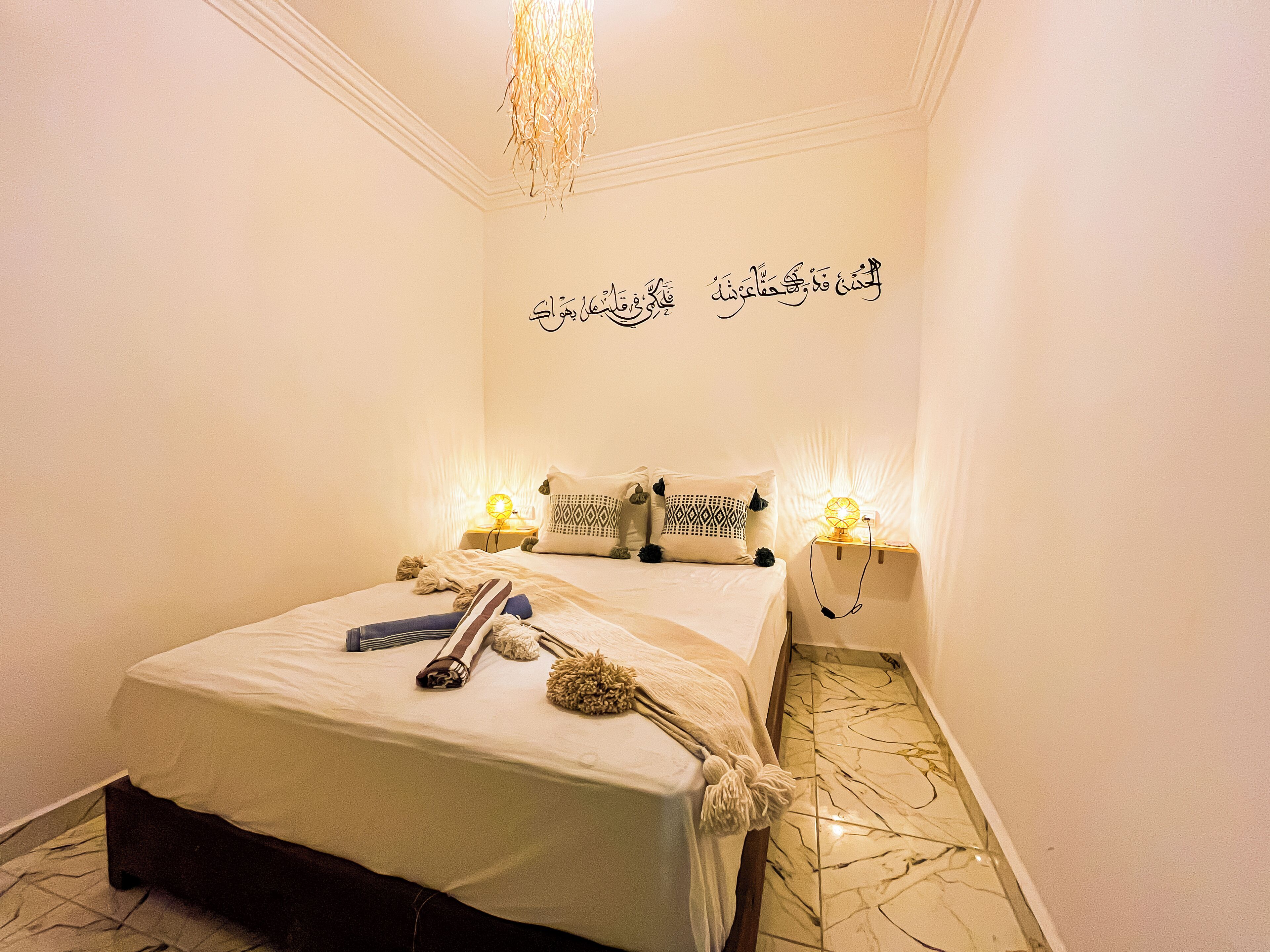 A neatly made bed with a white bedspread, two pillows, and a decorative wall with Arabic calligraphy.