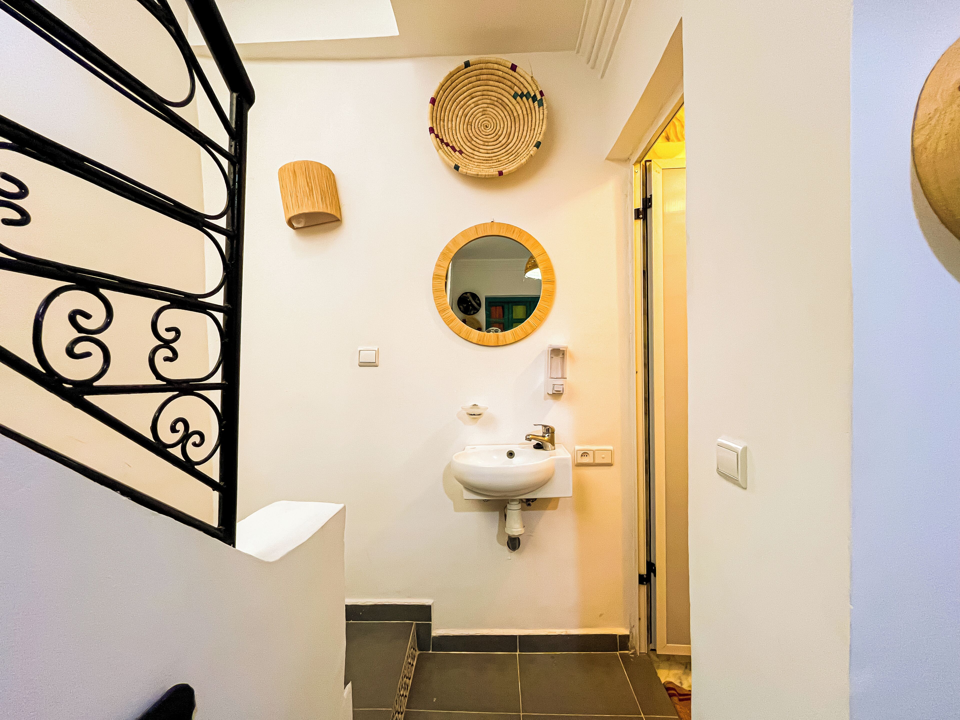 A narrow hallway with a white sink, a round mirror, and a woven wall hanging.