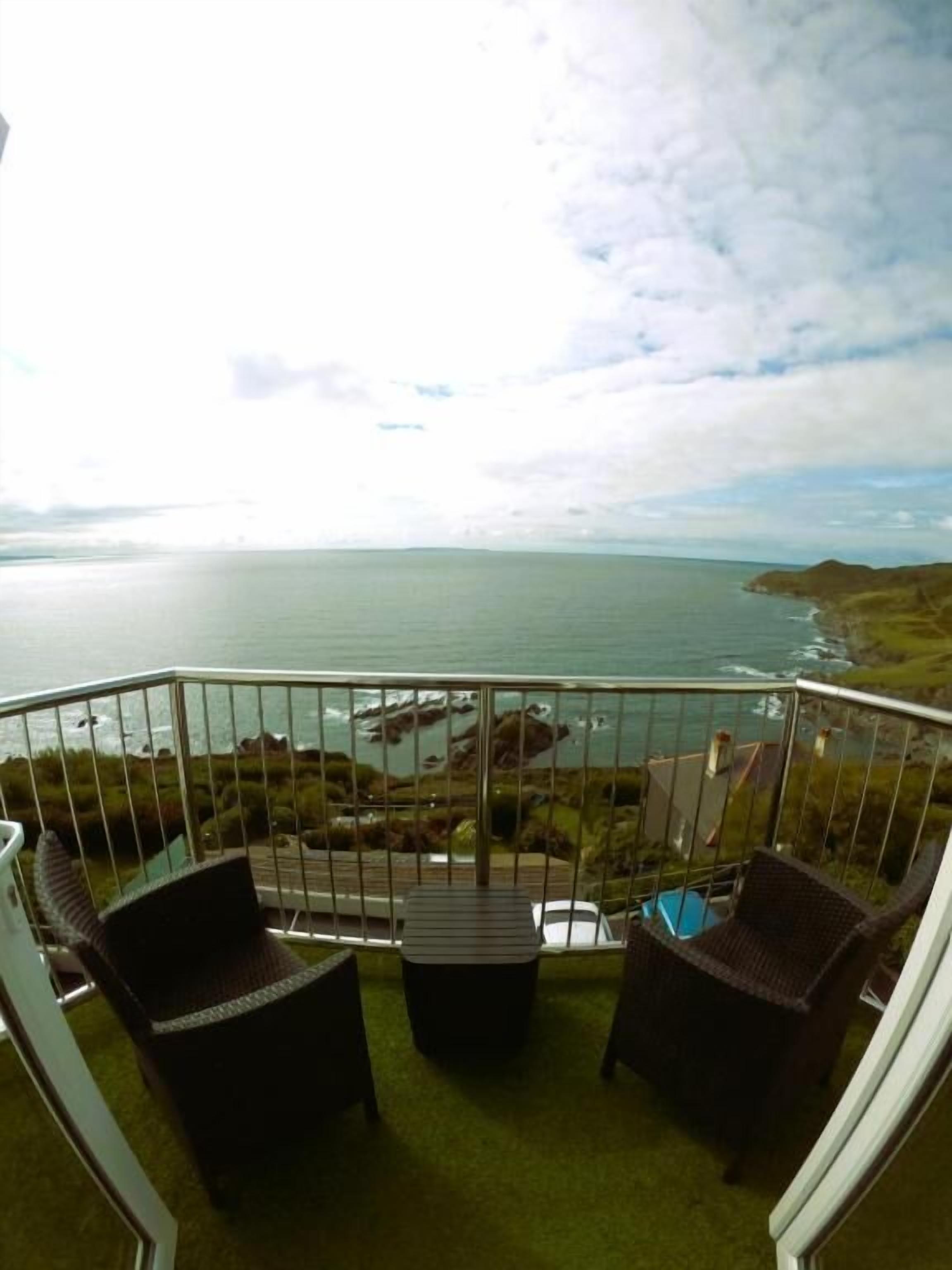 A balcony with two wicker chairs and a small table, overlooking a coastal view with the sea and distant land.