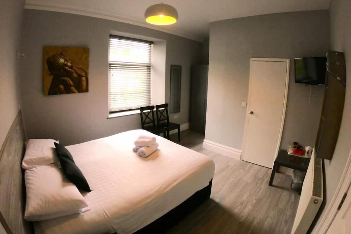 A bedroom with a bed, a bedside table, a chair, a window with blinds, a painting on the wall, and a door.
