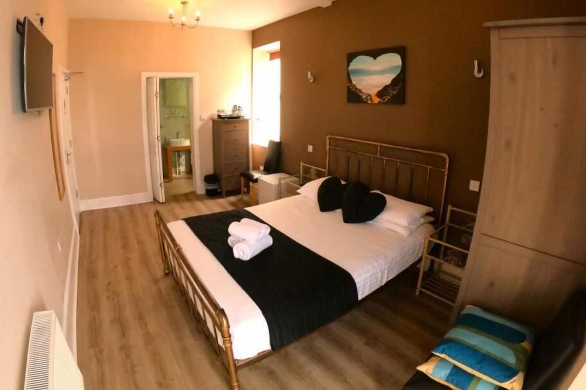 A bedroom with a bed, bedside tables, a dresser, a TV, and a door leading to a bathroom.