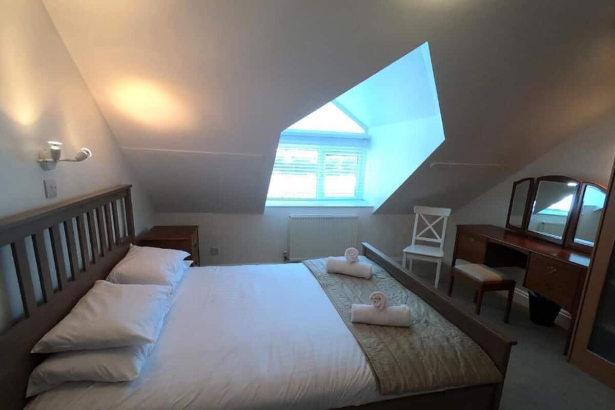 A bedroom with a skylight, a bed with pillows, a desk with a chair, and a radiator.