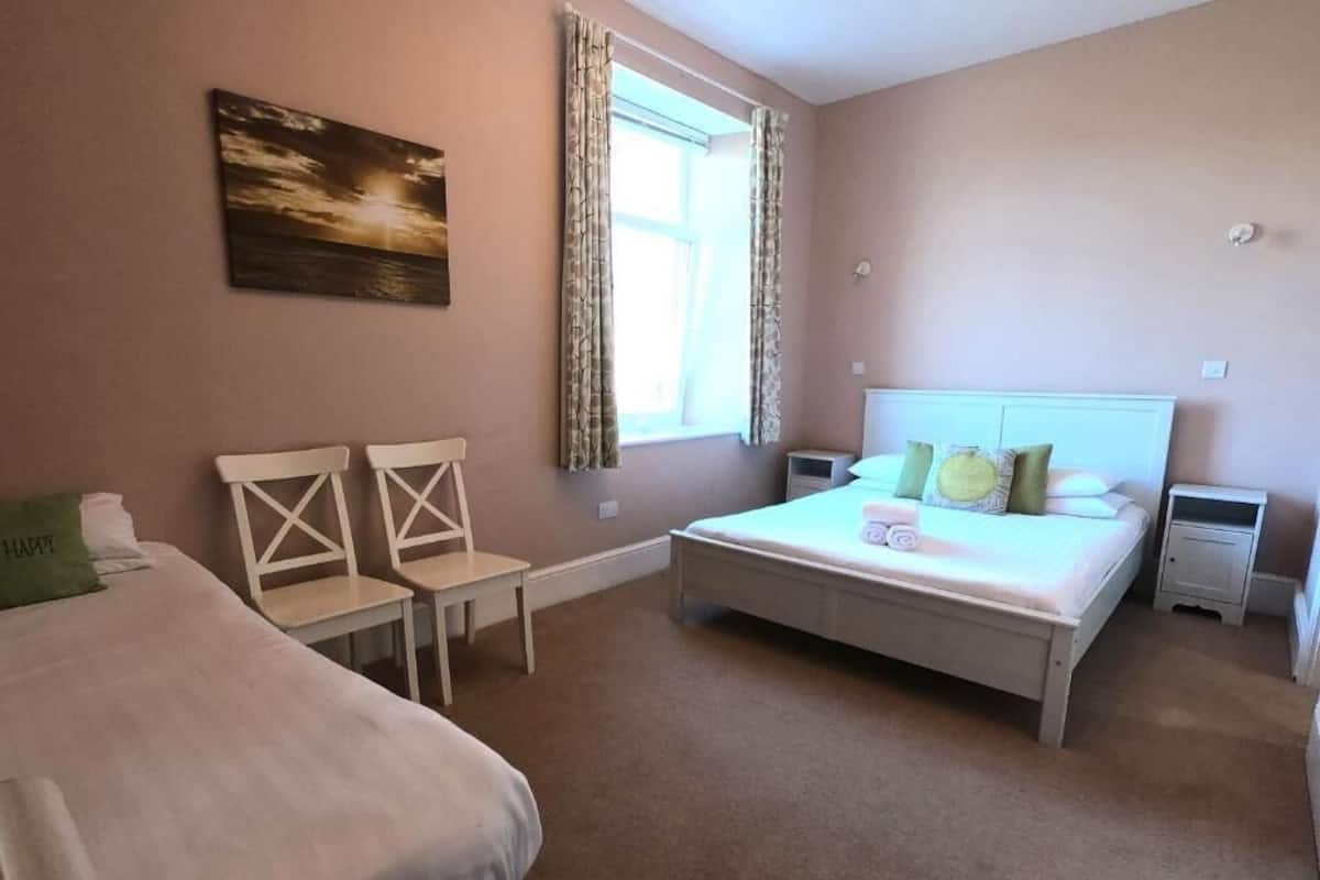 A bedroom with a bed, two chairs, a nightstand, a bedside table, and a window with curtains.
